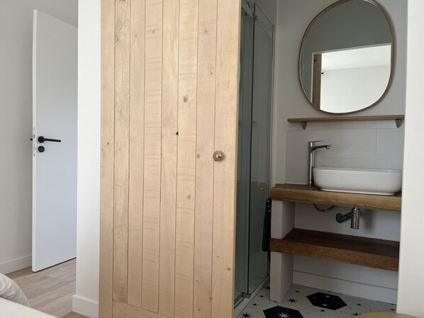 Ein Badezimmer mit Holztür, Duschkabine aus Glas, weißem Waschbecken mit Holzablage und einem Spiegel an der Wand.
