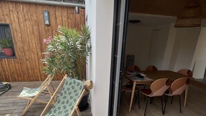 Een houten terras met een tafel en stoelen, een plant in pot en een schuifdeur naar een binnenruimte.