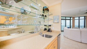 A modern kitchen with a glass display shelf, stainless steel sink, and a large window offering a view of the outdoors.