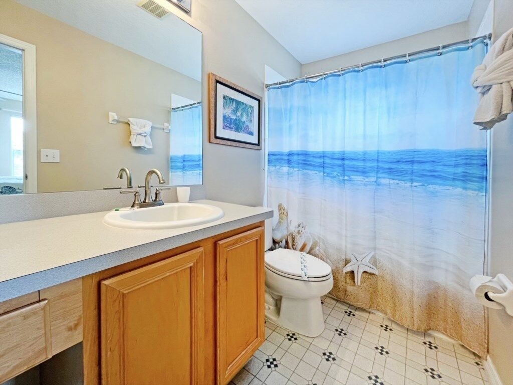 A bathroom with a large mirror, two sinks, a toilet, and a shower curtain depicting a beach scene.