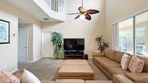 A spacious living room with a large sectional sofa, a flat-screen TV, a ceiling fan, and a large window providing natural light. - Sunnyside Villa 6 Bedroom Home by RedAwning (Kissimmee)