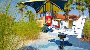 A playground with a swing set and a slide, surrounded by palm trees and a building in the background. - Sunnyside Villa 6 Bedroom Home by RedAwning (Kissimmee)
