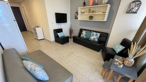 Un salon avec un canapé gris, un fauteuil noir, une petite table ornée d’objets décoratifs, un téléviseur à écran plat et une étagère contenant divers objets.
