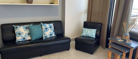 A living room with a black sofa, a matching armchair, a small wooden table, and a shelf with decorative items.