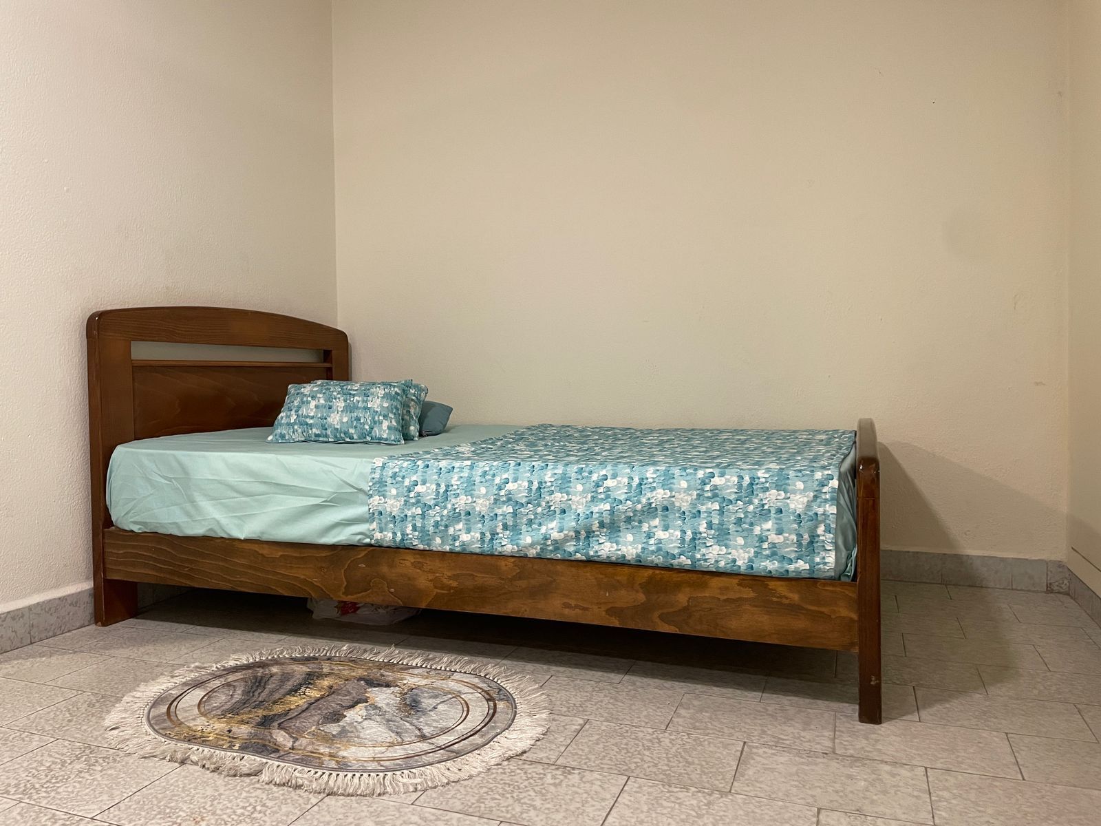 A single bed with a wooden headboard, a patterned bedspread, and a circular rug on a tiled floor.