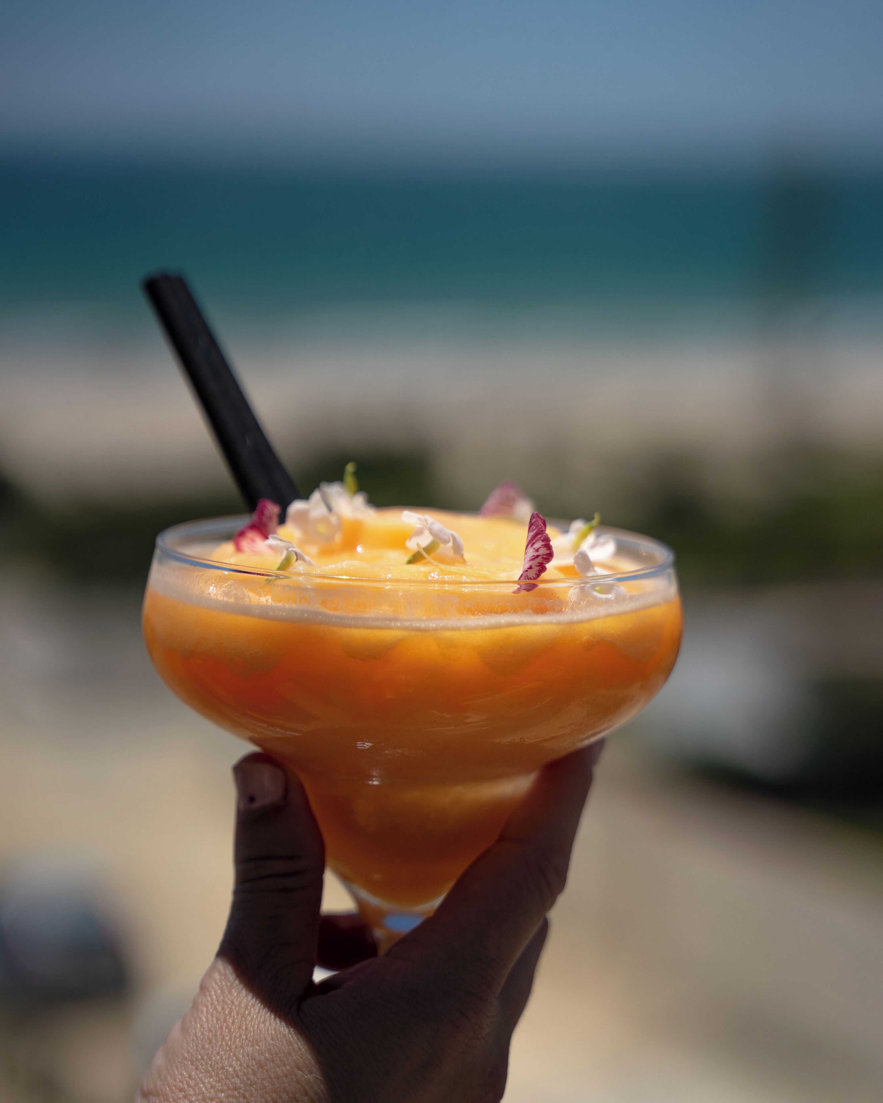 A hand holding a cocktail with a straw, garnished with flowers, against a beach backdrop.