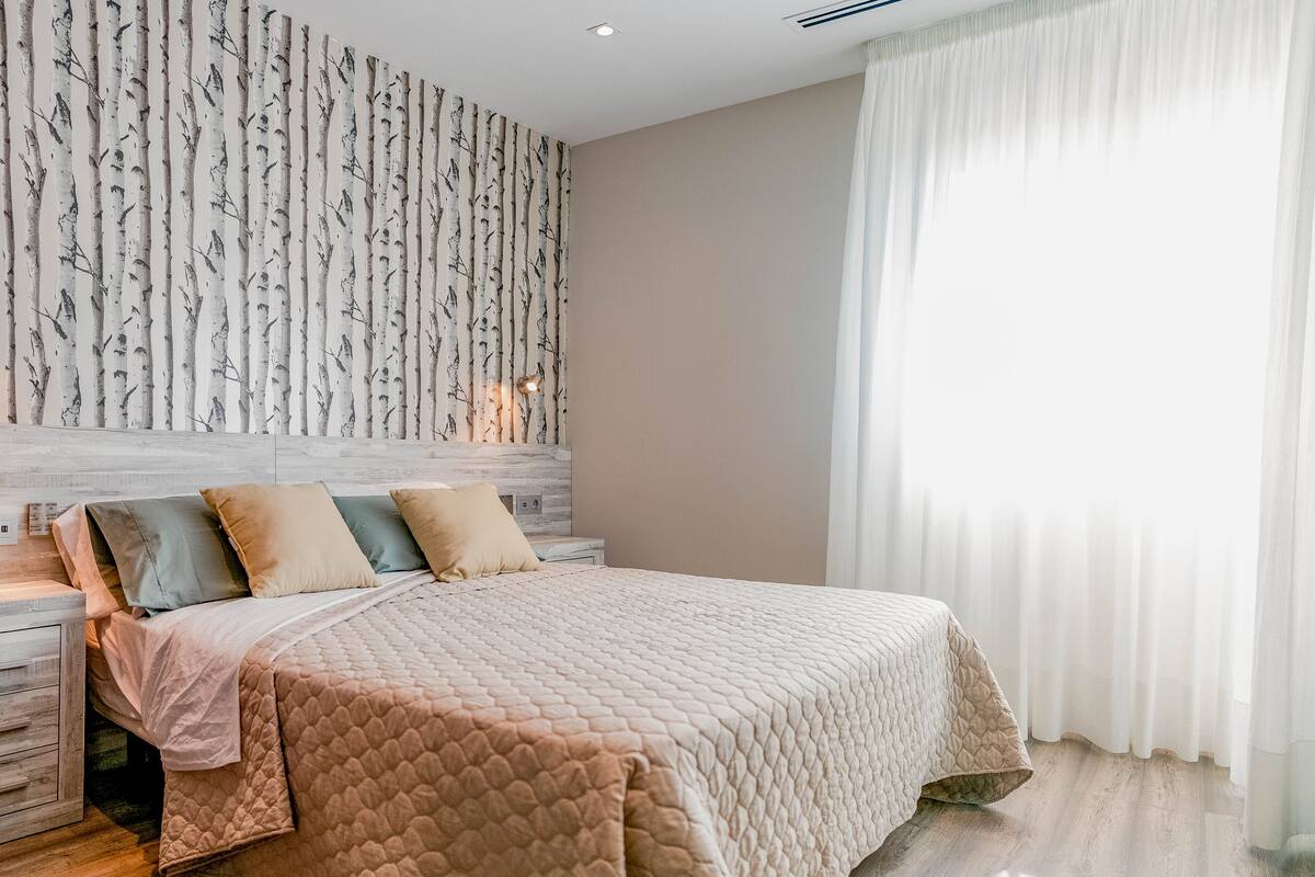 Une chambre à coucher avec un lit, une table de chevet, un mur orné d’un motif d’arbre et une fenêtre avec des rideaux.