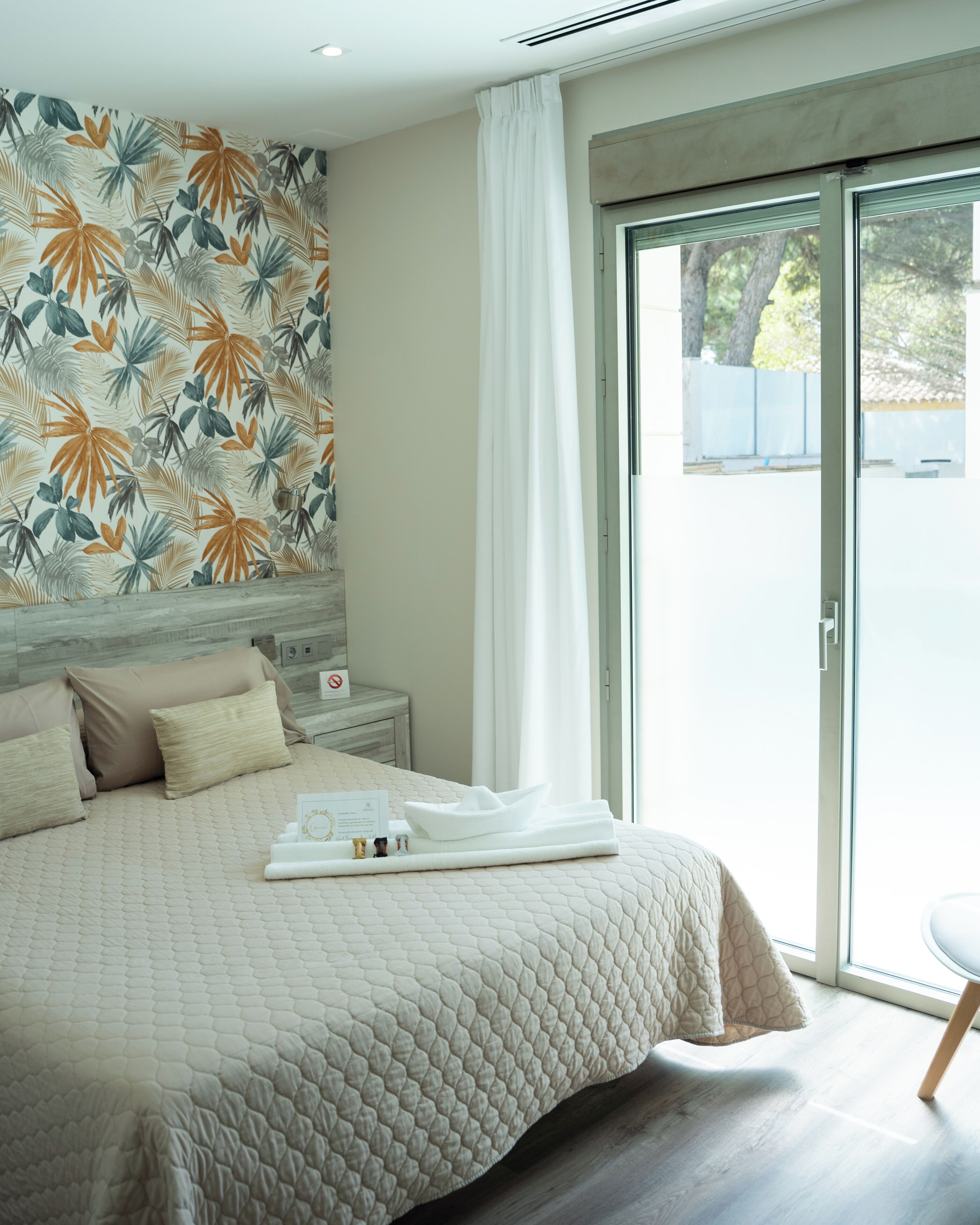A modern hotel room with a large bed, a nightstand, a window with curtains, and a tropical leaf-patterned wall.