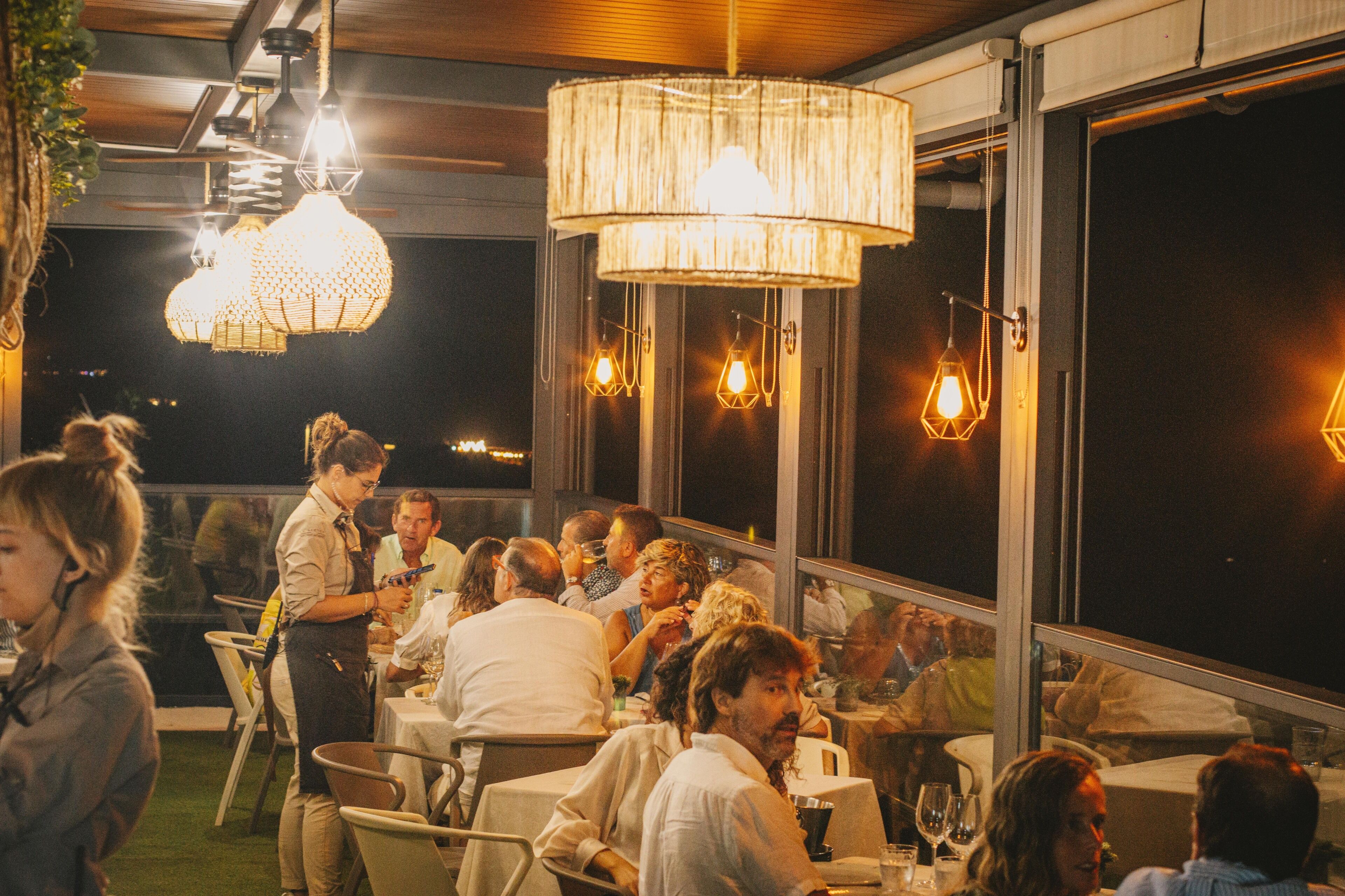 An outdoor dining area with tables and chairs, large windows, and hanging pendant lights.