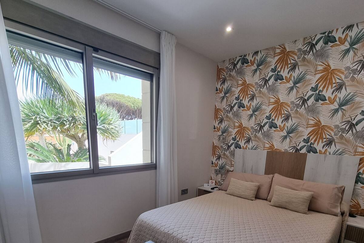 Une chambre avec un grand lit, une table basse en bois, une fenêtre avec des rideaux et un papier peint aux motifs tropicaux.