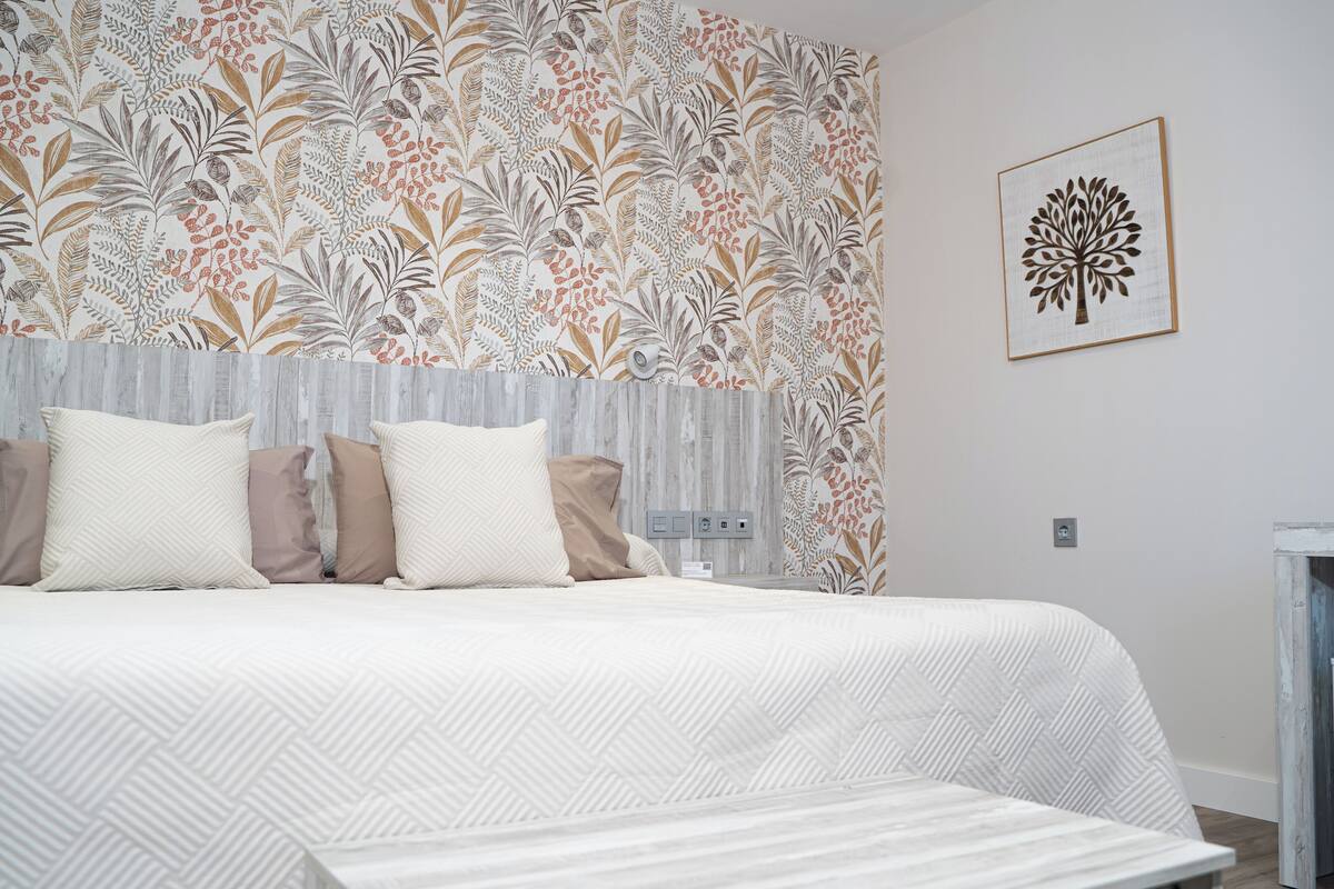 Une chambre à coucher avec un lit, des oreillers, une tête de lit, un mur orné d’un motif floral et un tableau encadré.