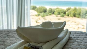 A neatly made bed with folded towels and a pair of slippers, a window with a view of the beach and ocean, and a package of toiletries.