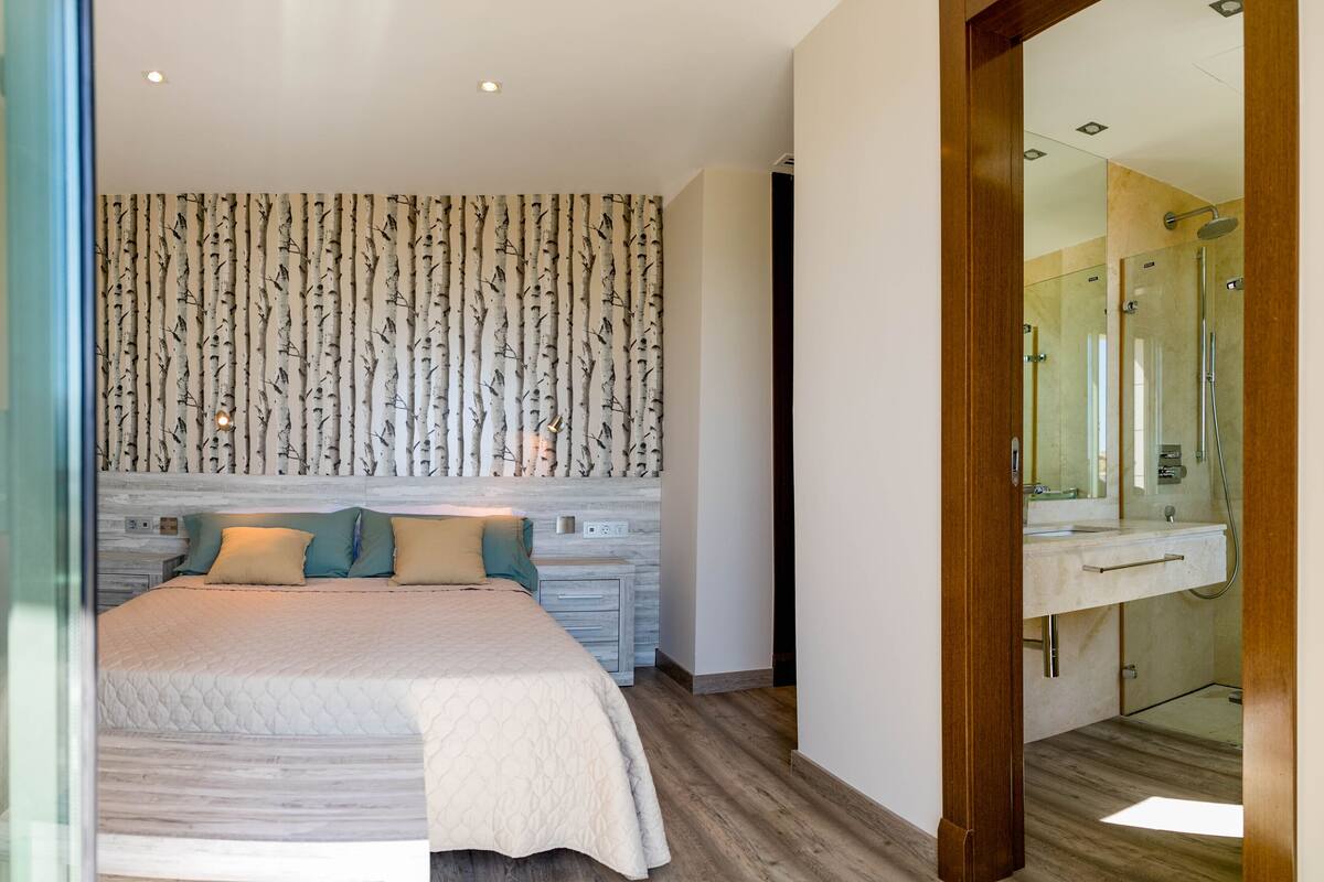 Une chambre moderne avec un grand lit, une tête de lit en bois et un mur orné d’un motif représentant un bouleau.