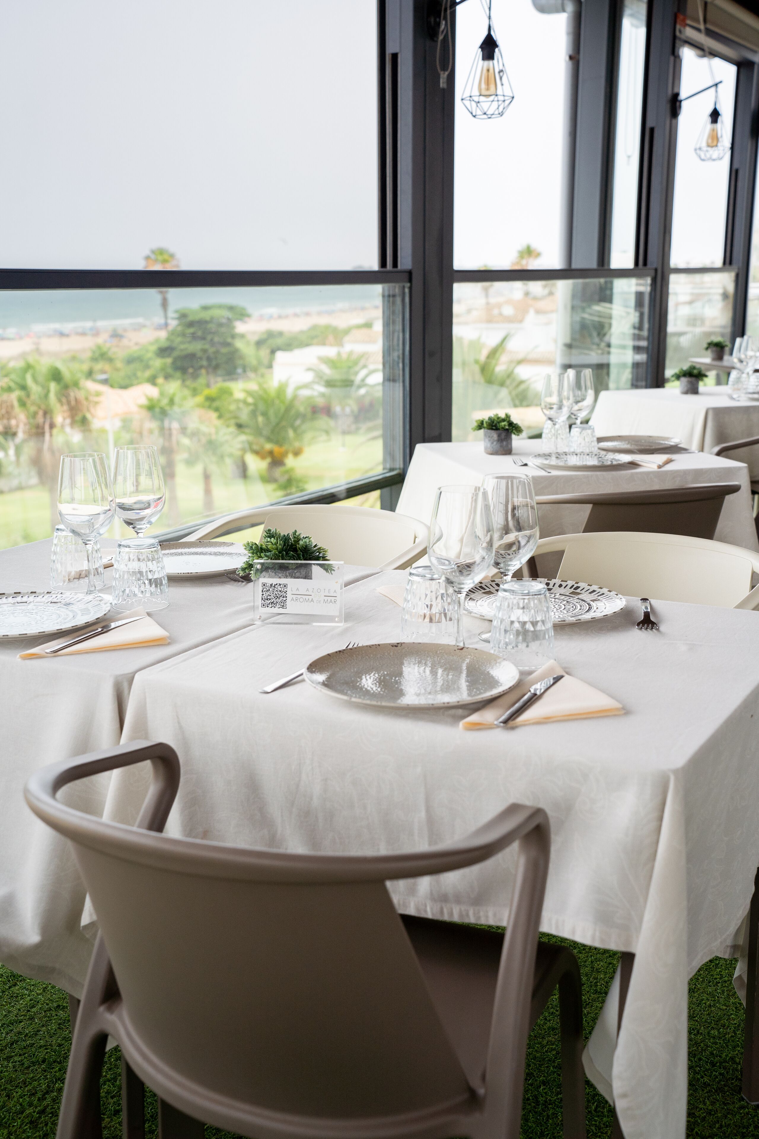 Comedor con mesas dispuestas para dos, con cristalería y platos transparentes, y vistas a la playa y palmeras a través de amplios ventanales.
