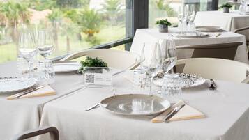 Comedor con mesas dispuestas para dos, con cristalería y platos transparentes, y vistas a la playa y palmeras a través de amplios ventanales.