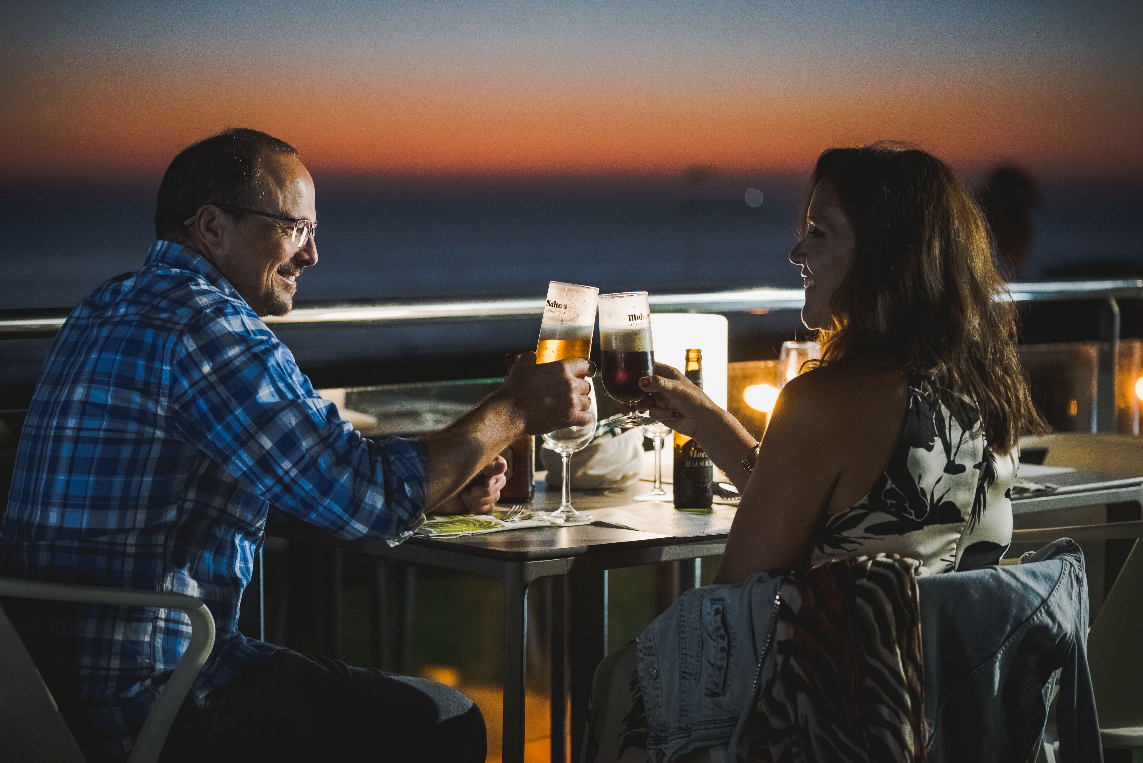 Two people toasting with drinks at a table with a view of the sunset.