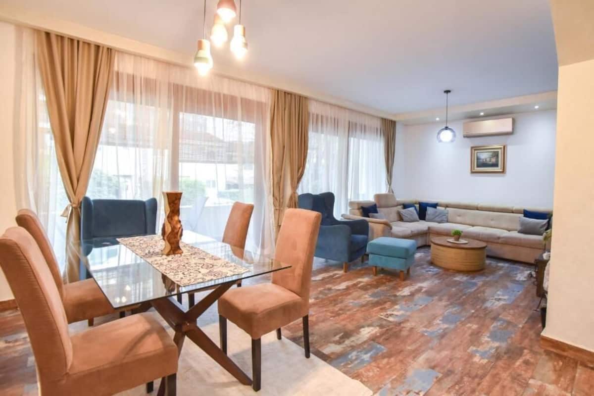 A modern living room with a glass dining table, comfortable seating, and a large window with sheer curtains.
