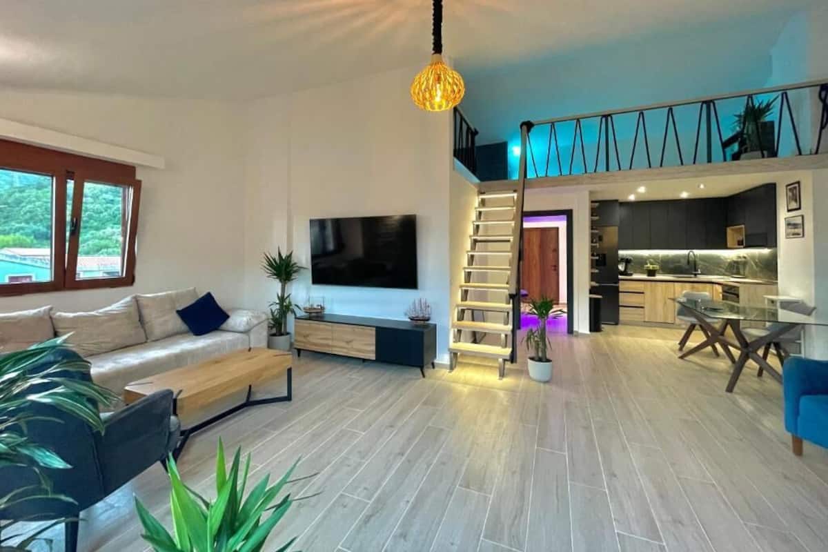A spacious living area with a sofa, coffee table, and TV. A staircase leads to a kitchen area with modern appliances.
