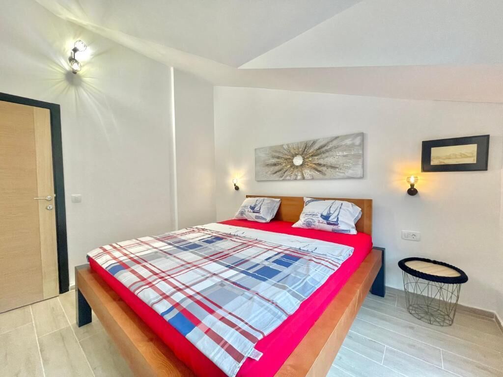 A bedroom with a bed, a wooden headboard, a wall-mounted artwork, a small round table, and a wall-mounted light fixture.