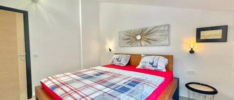 A bedroom with a bed, a wooden headboard, a wall-mounted artwork, a small round table, and a wall-mounted light fixture.