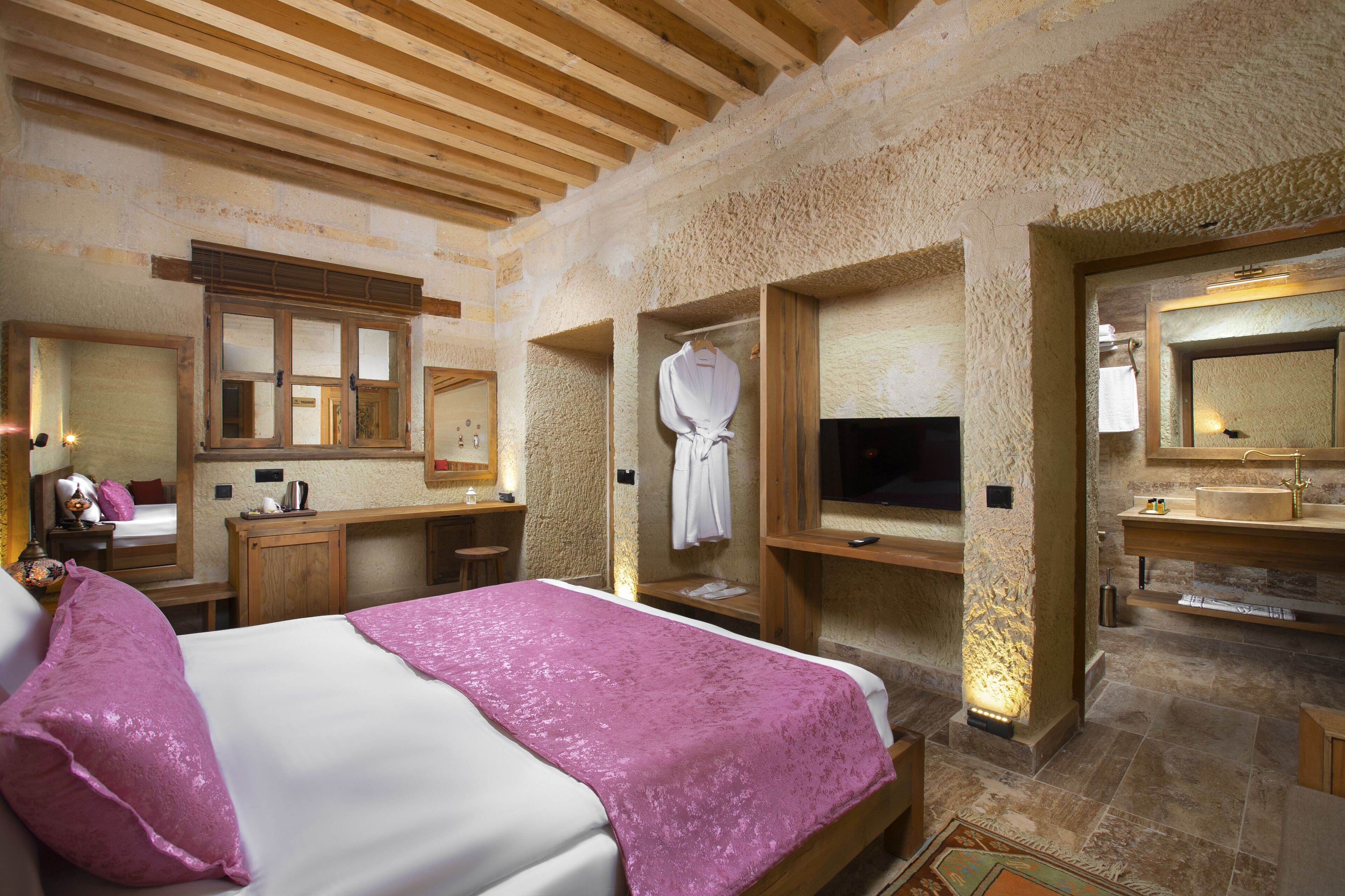 A bedroom with a large bed, wooden beams, stone walls, a wooden dresser, a TV, and a bathroom with a sink and mirror.