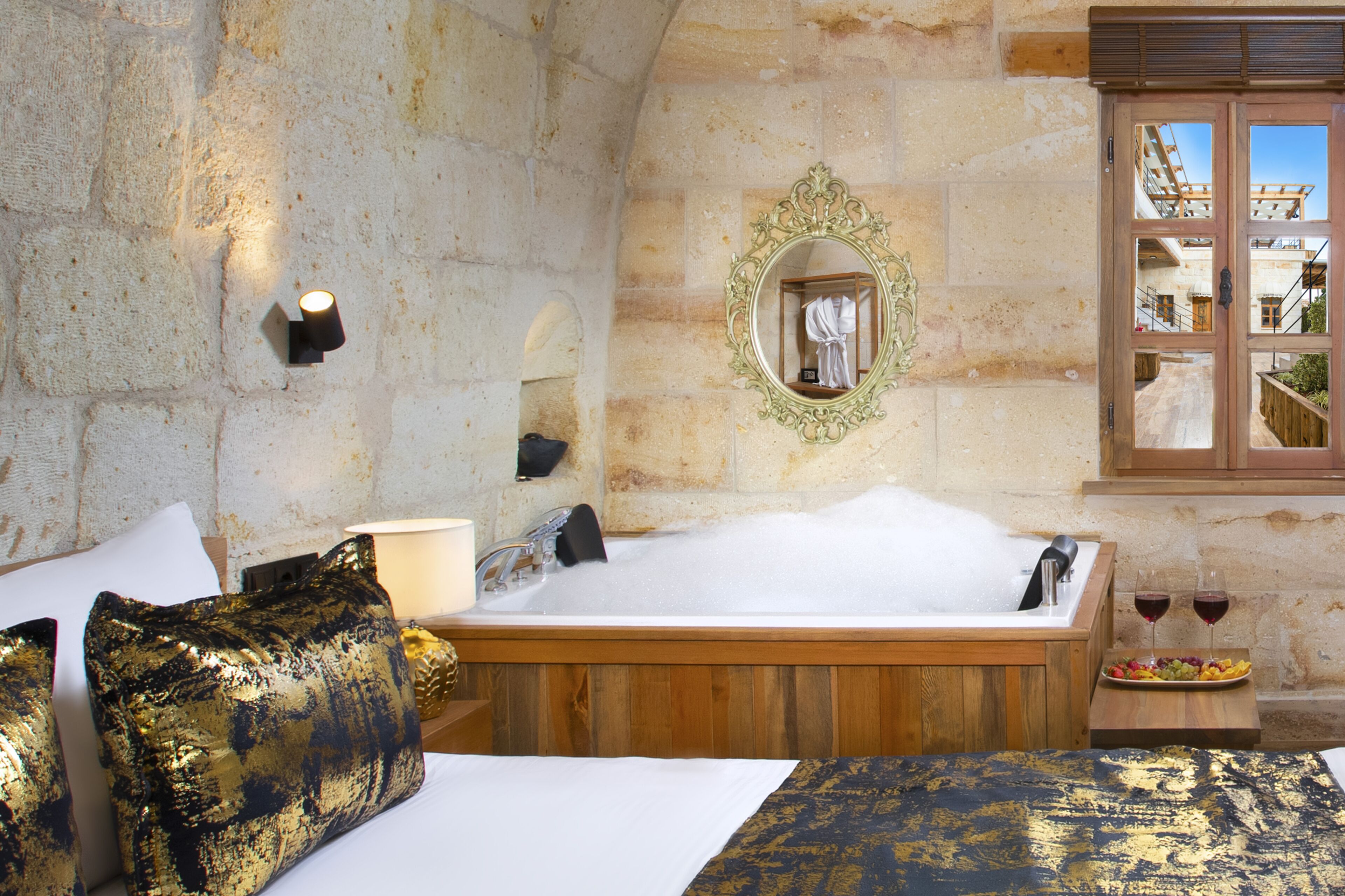 Una habitación con una bañera de madera, una cama con almohadas de diseño dorado y negro, un espejo y una bandeja de frutas con dos copas de vino tinto.