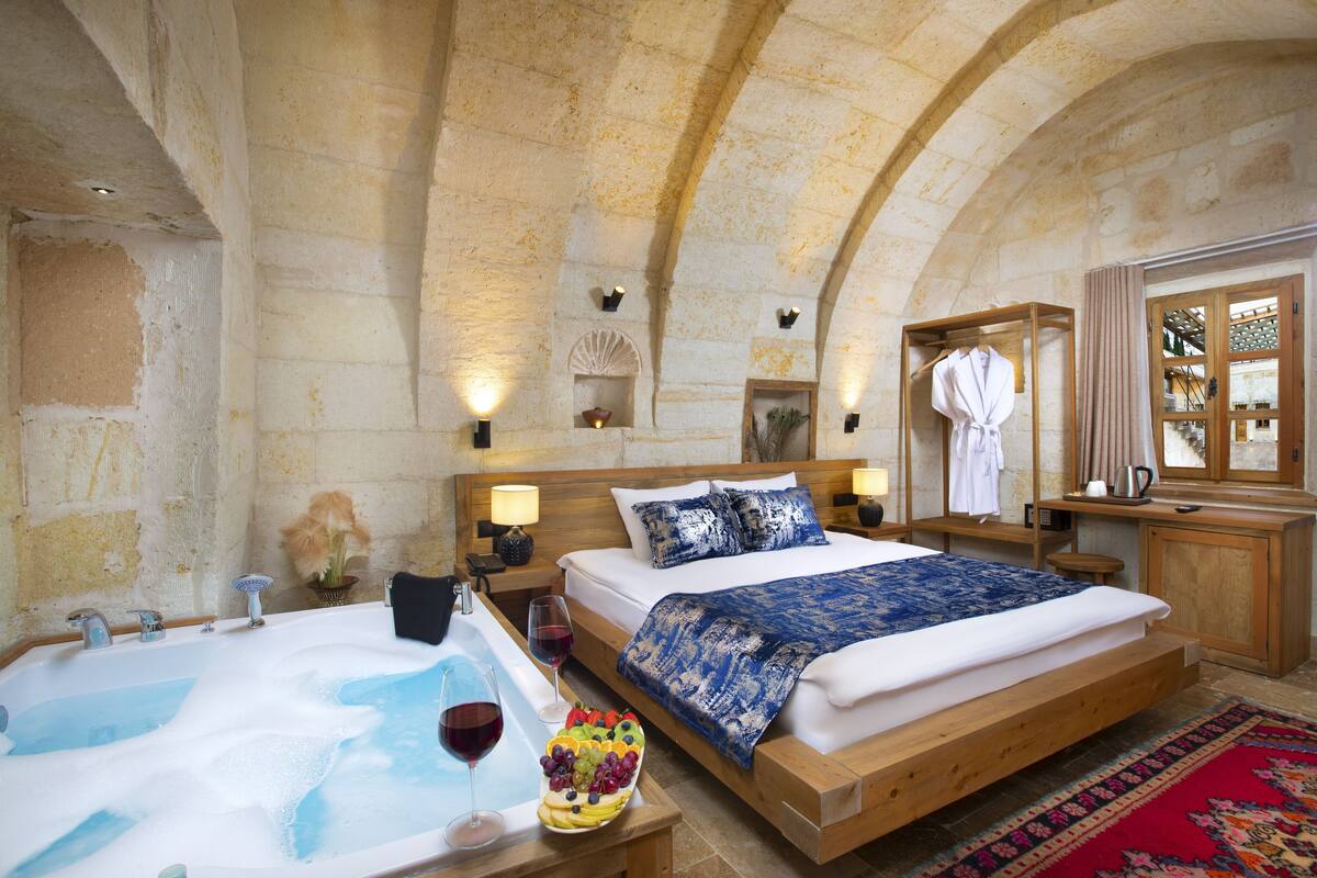A room with a large bed, a wooden headboard, a bathtub with bubbles, and a fruit bowl on the side.