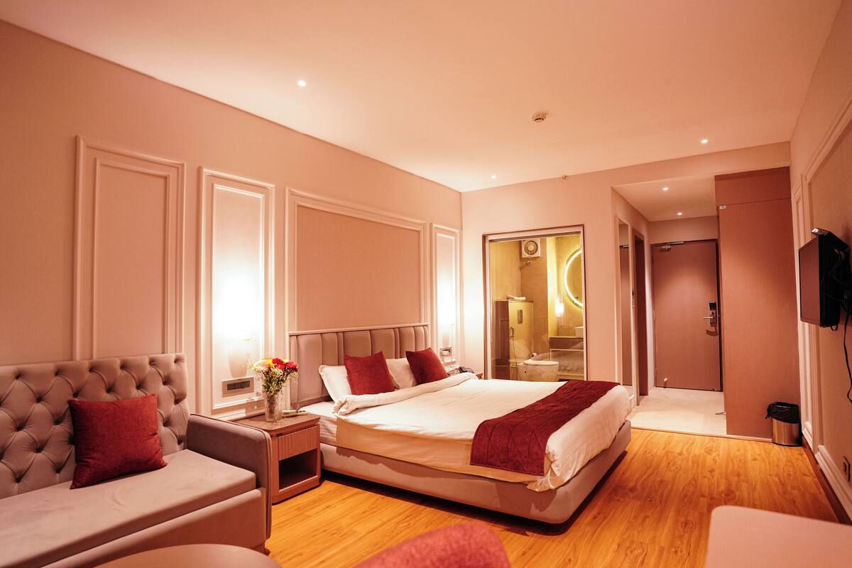 A modern hotel room with a large bed, a sofa, a bedside table with a vase of flowers, and a mirror.