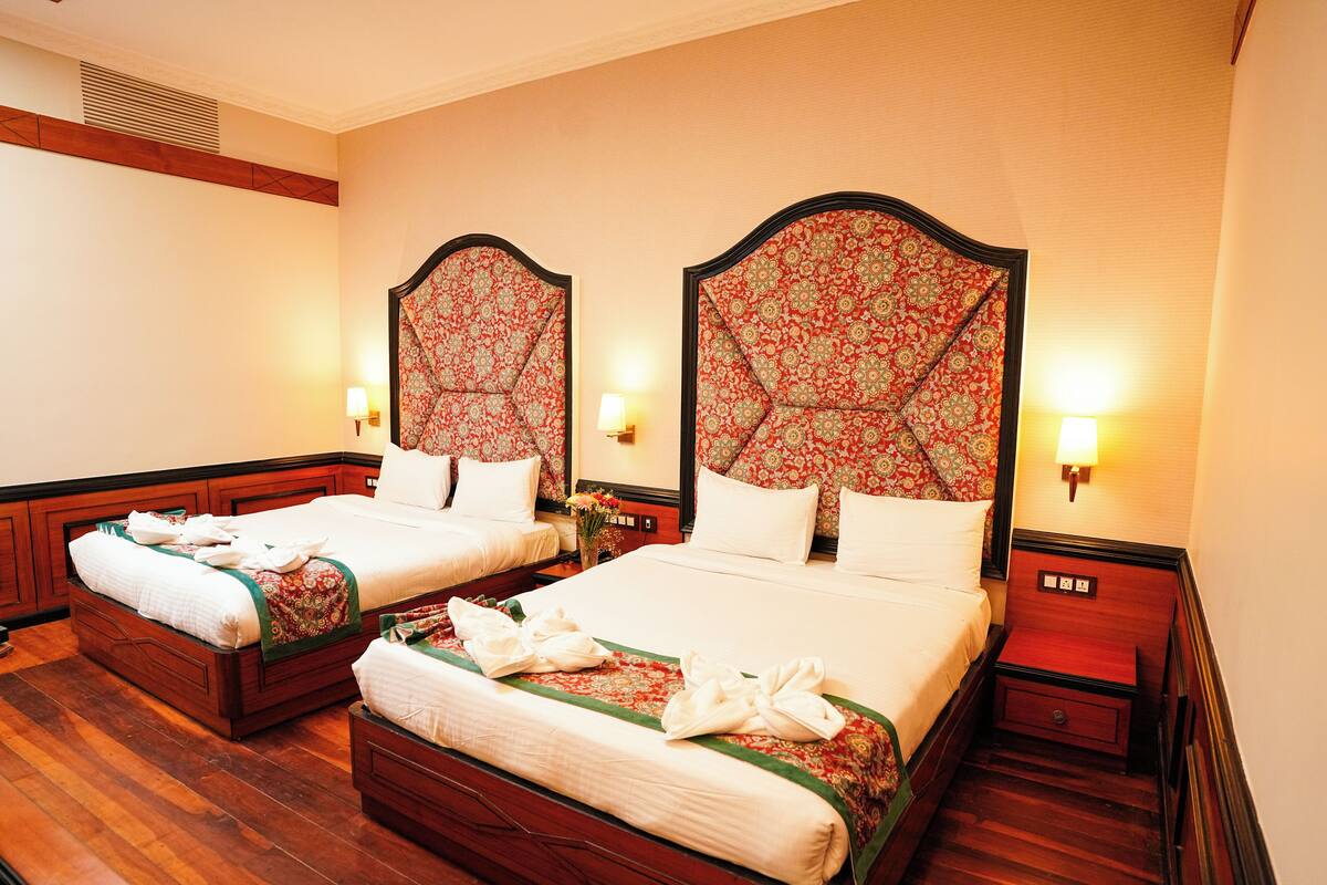A hotel room with two beds, each with a wooden headboard and floral-patterned bedding. There are bedside tables with lamps and a small drawer.