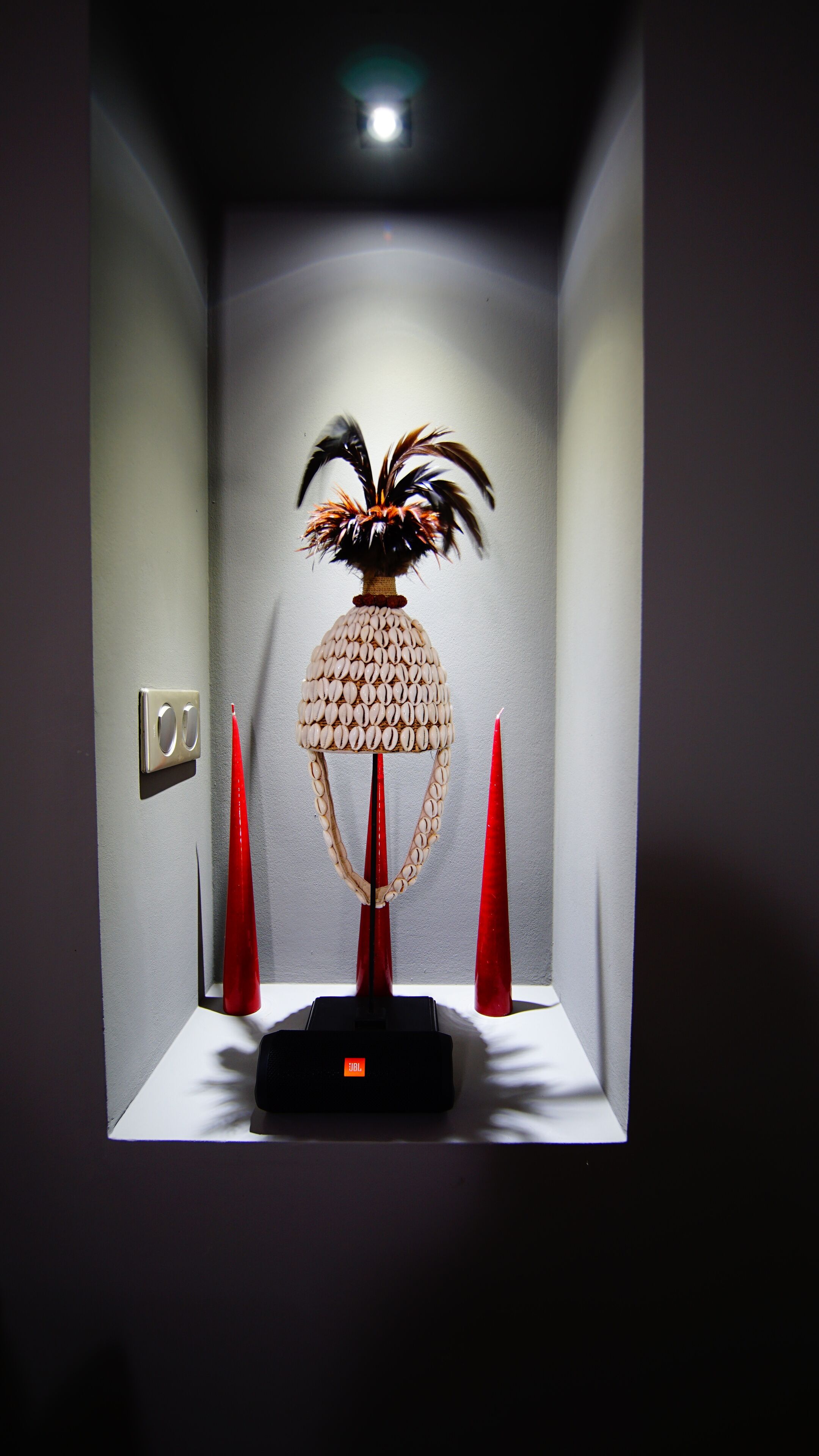 Une pièce avec une lampe décorative posée sur un piédestal, un interrupteur fixé au mur et deux bougies rouges.