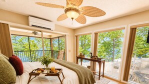 Un dormitorio con cama, ventilador de techo, ventana con vista a árboles y una mesita con bandeja de comida.