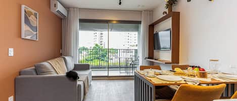 A modern living room with a grey sofa, a dining table set for two, and a TV mounted on the wall.