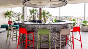 A modern bar area with a circular design, surrounded by green and red chairs, and large windows offering a city view. - Charlie For You Moema (São Paulo)