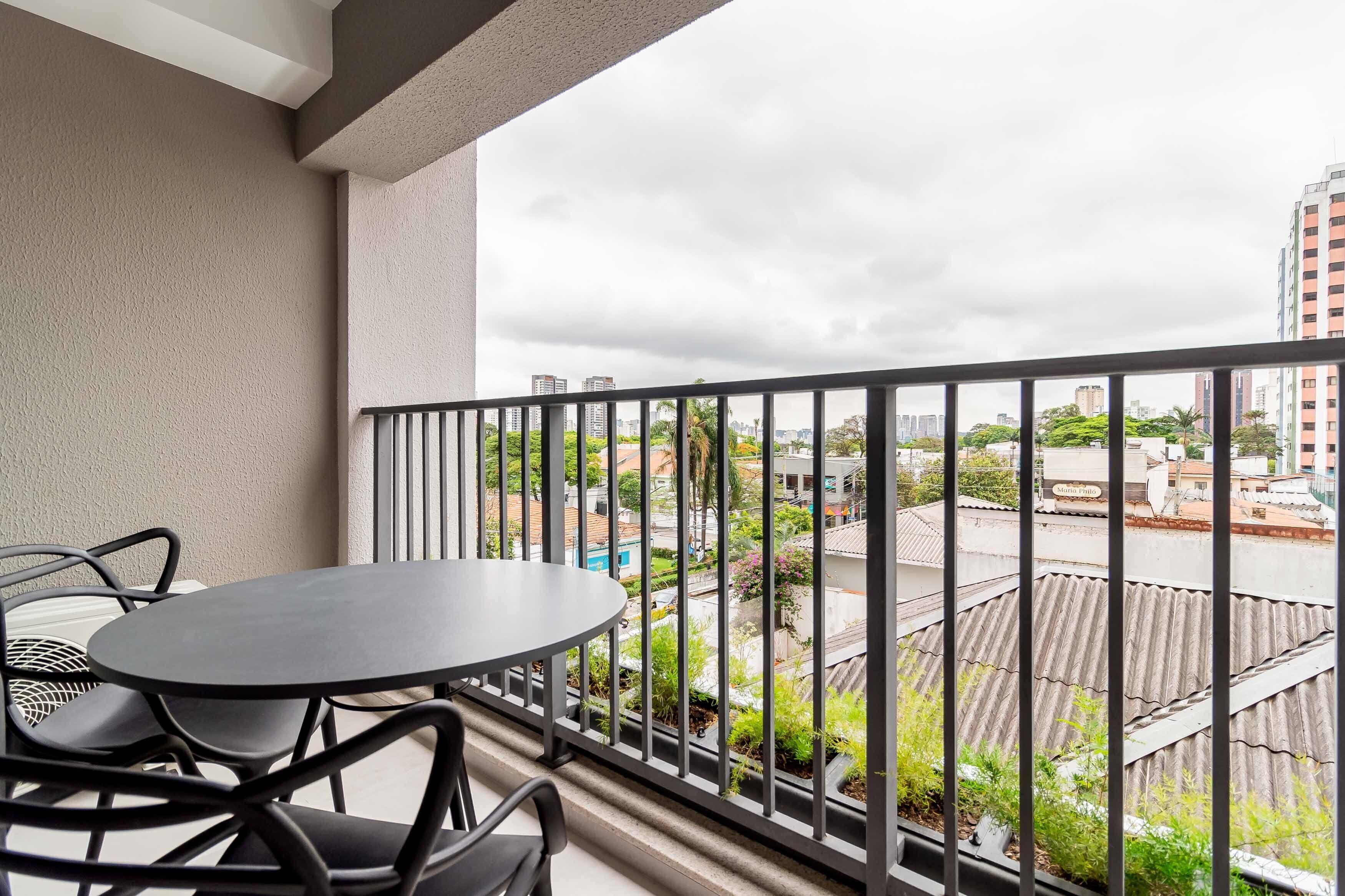 A balcony with a round table and chairs, a view of a cityscape with buildings and greenery, and a cloudy sky.