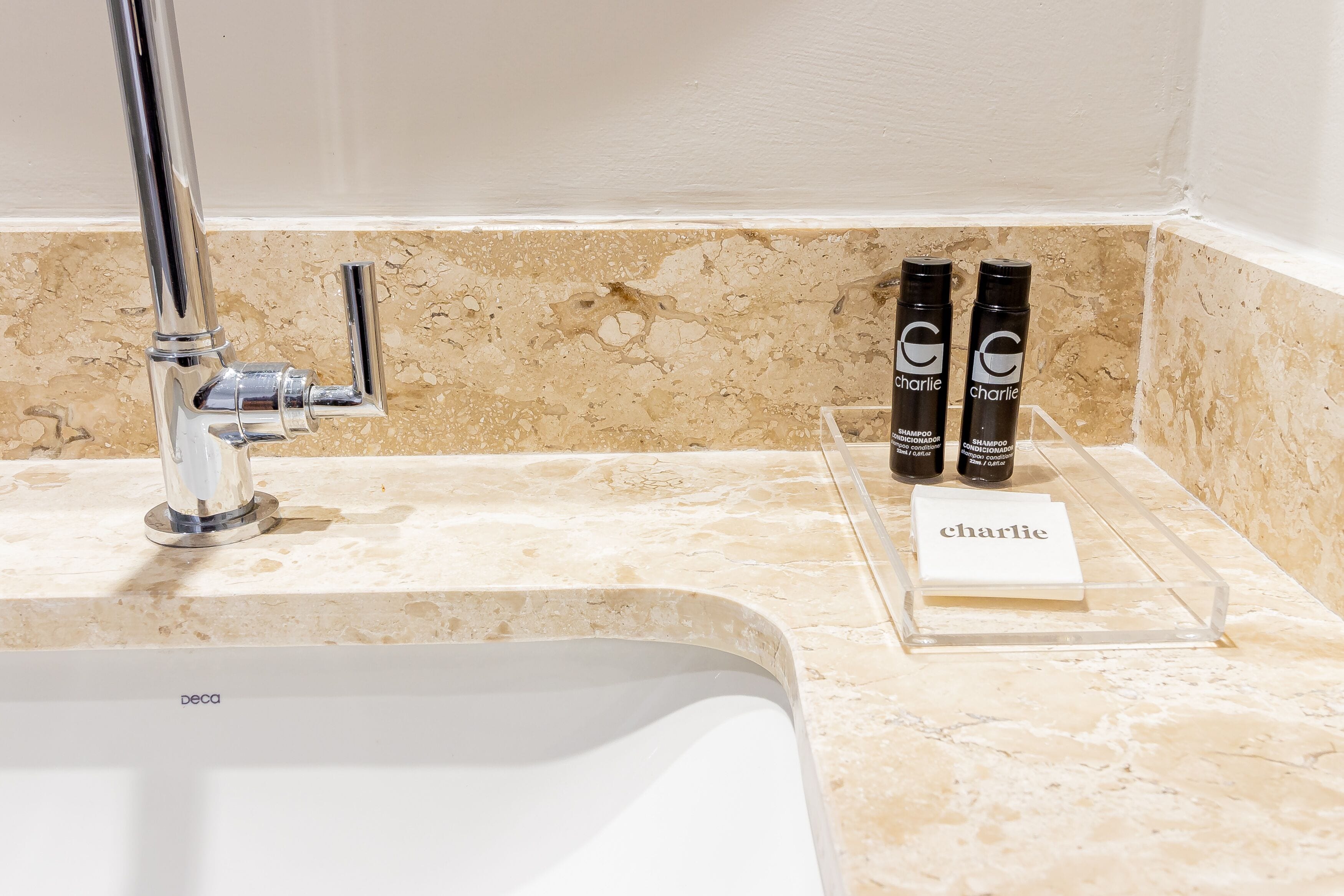 A bathroom with a white sink, chrome faucet, and two bottles of shampoo labeled 'Charlie' on a clear shelf.