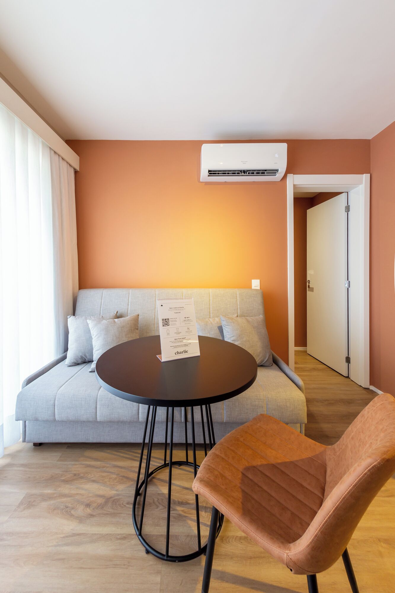 A modern living room with a grey sofa, a round table, and a single brown chair. An air conditioning unit is mounted on the wall above the sofa.