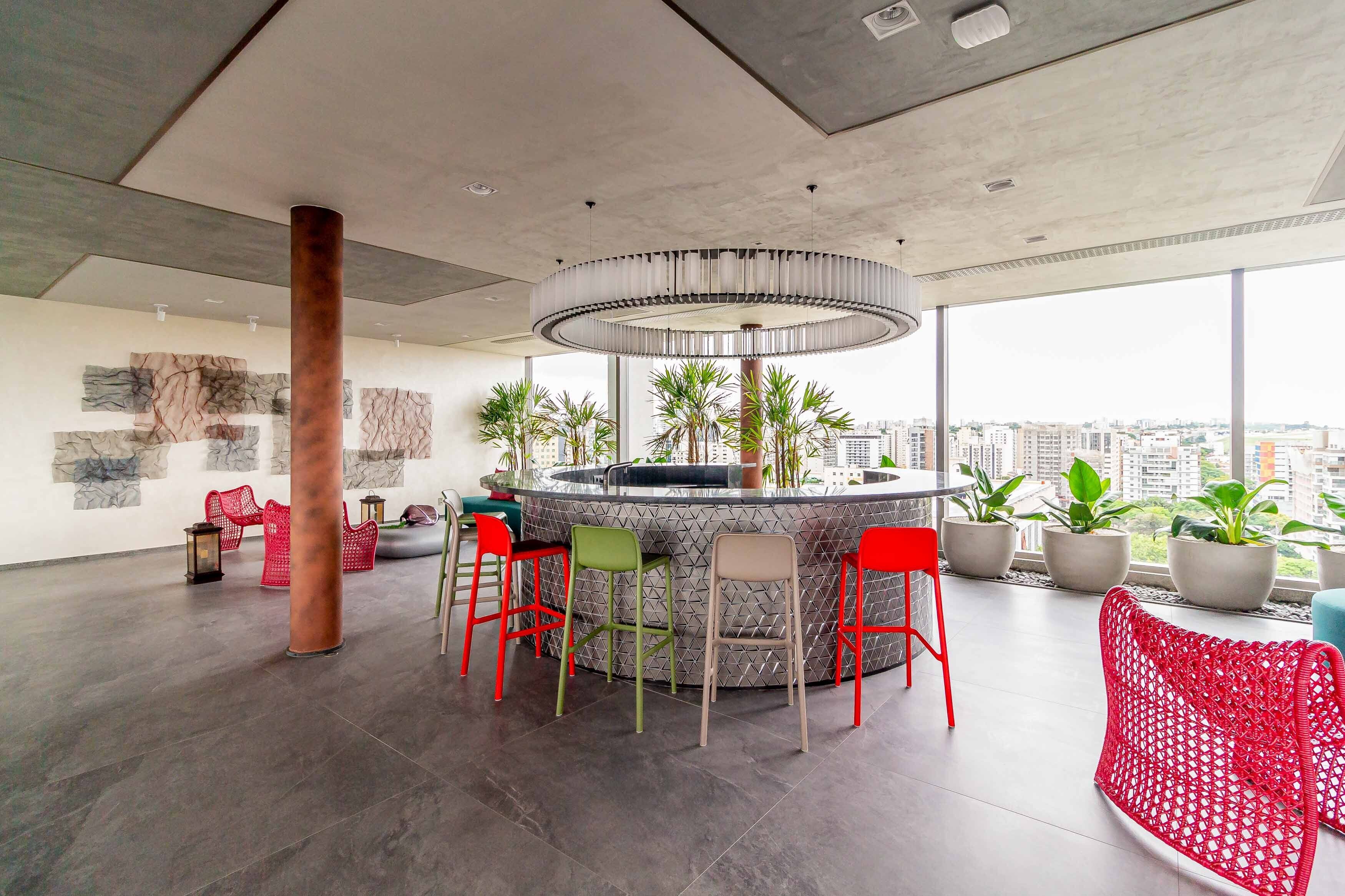 Un área de bar moderna con diseño circular, rodeada de sillas rojas y verdes, y grandes ventanales con vista a la ciudad.