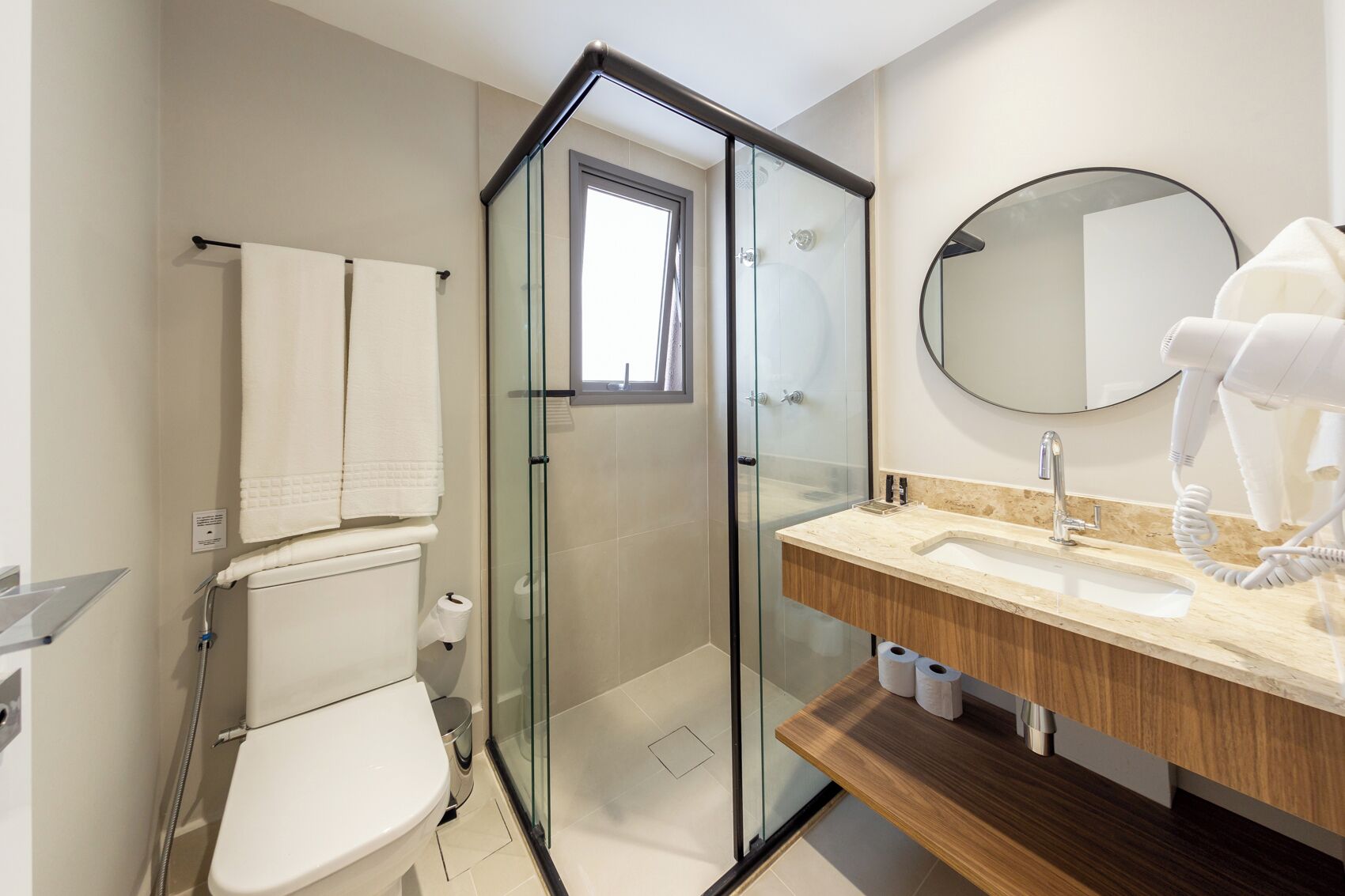 A modern bathroom with a glass-enclosed shower, a white toilet, a wooden vanity with a sink, and a round mirror.