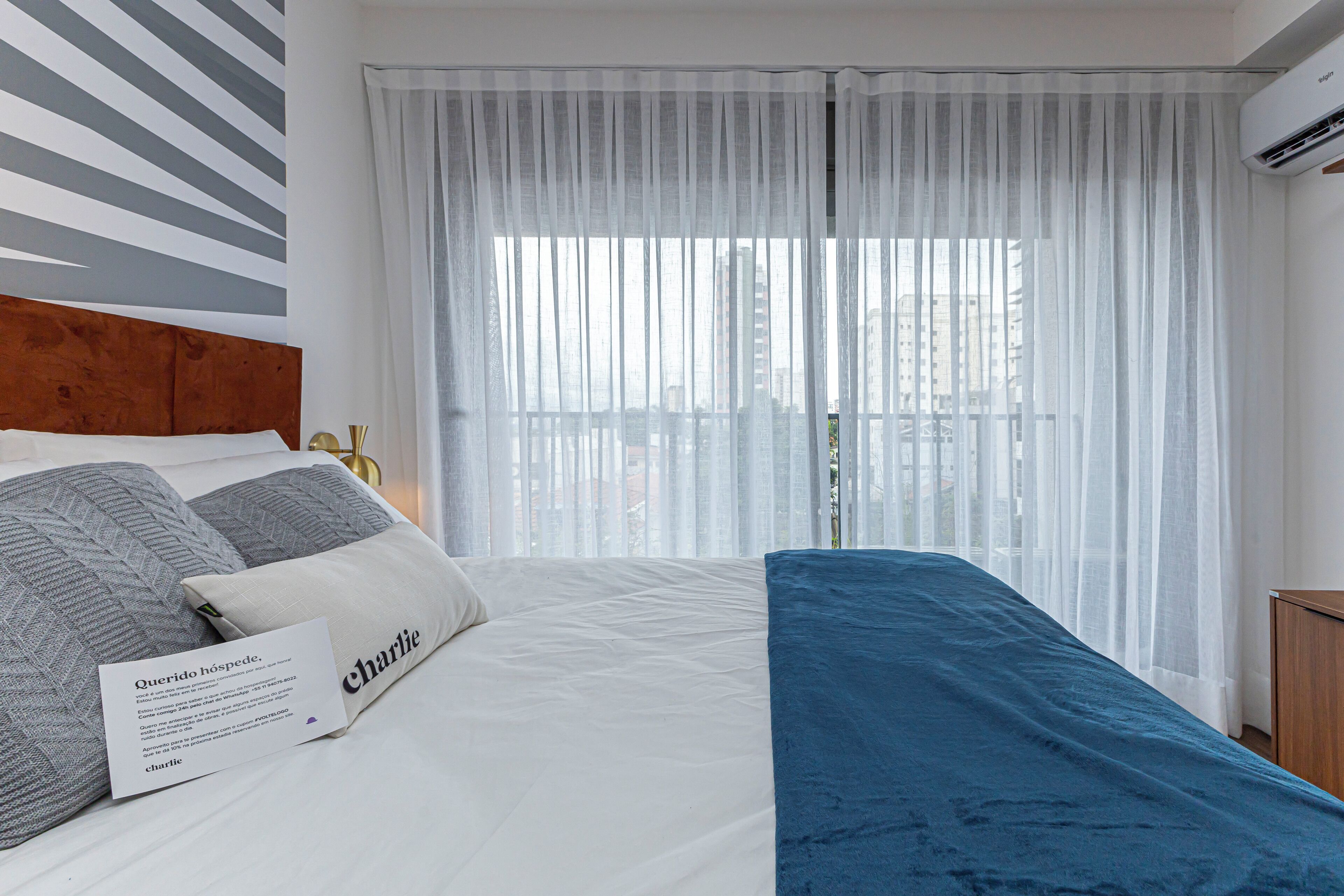 A hotel room with a bed, a blue sofa, a white curtain, and a view of buildings through the window.