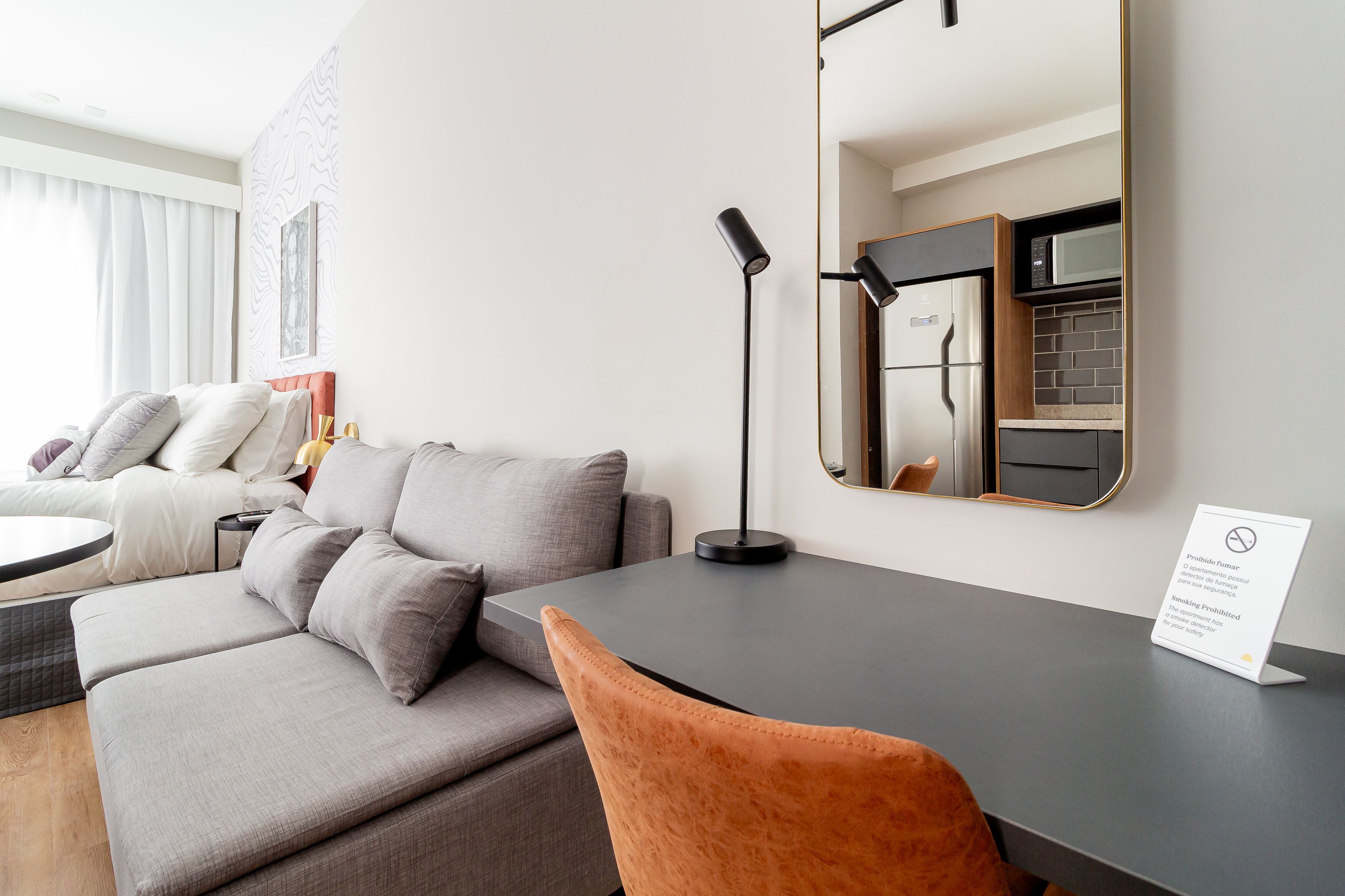 A modern hotel room with a grey sofa, a small round table, a black desk, a mirror, and a sign indicating smoking is prohibited.