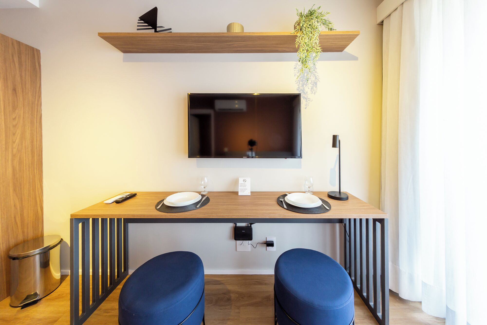 A modern dining area with a wooden table, two blue chairs, a TV mounted on the wall, and a hanging plant.
