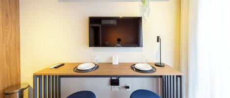 A modern dining area with a wooden table, two blue chairs, a TV mounted on the wall, and a hanging plant.