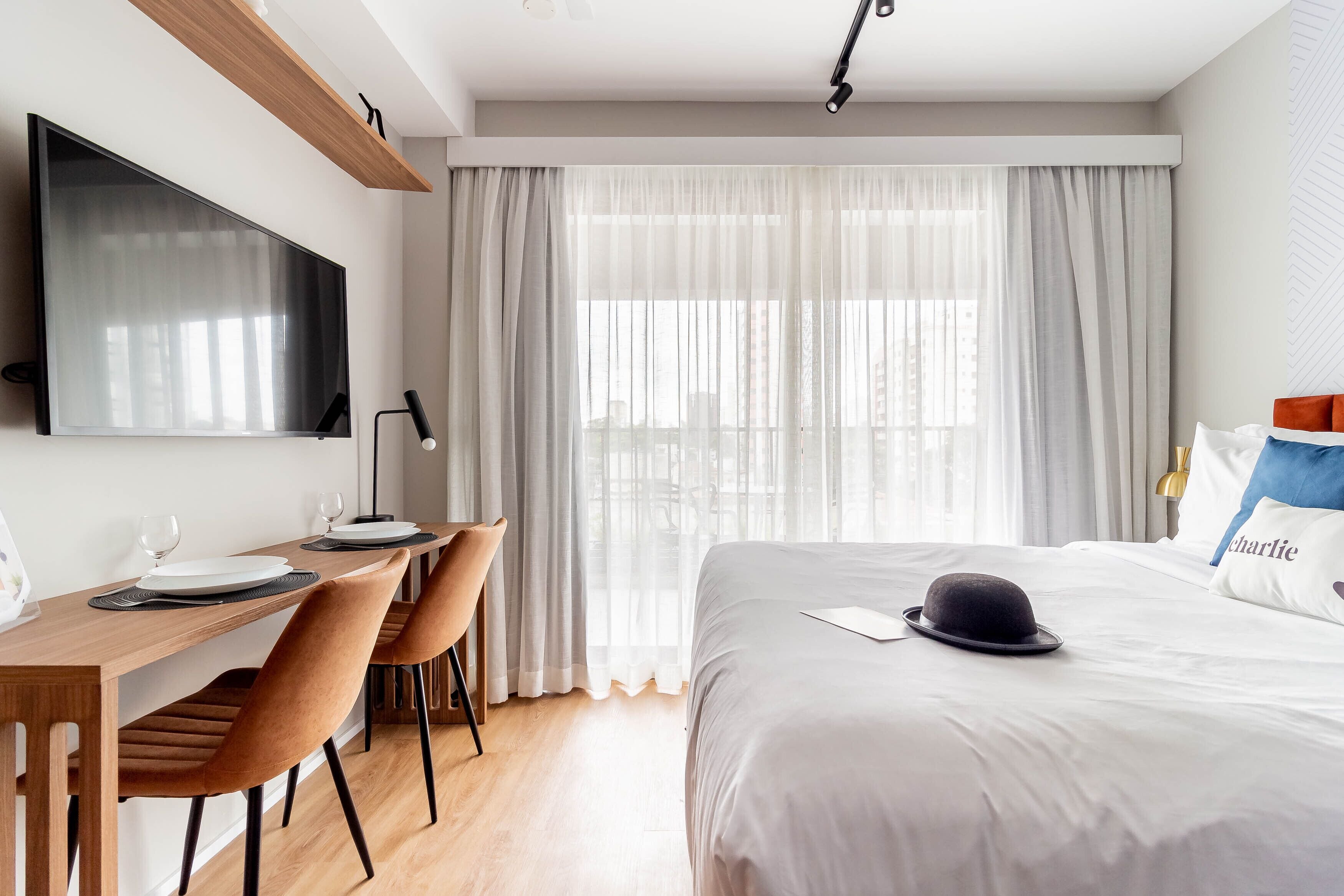 A modern hotel room with a large bed, a desk with chairs, a flat-screen TV, and a large window with sheer curtains.