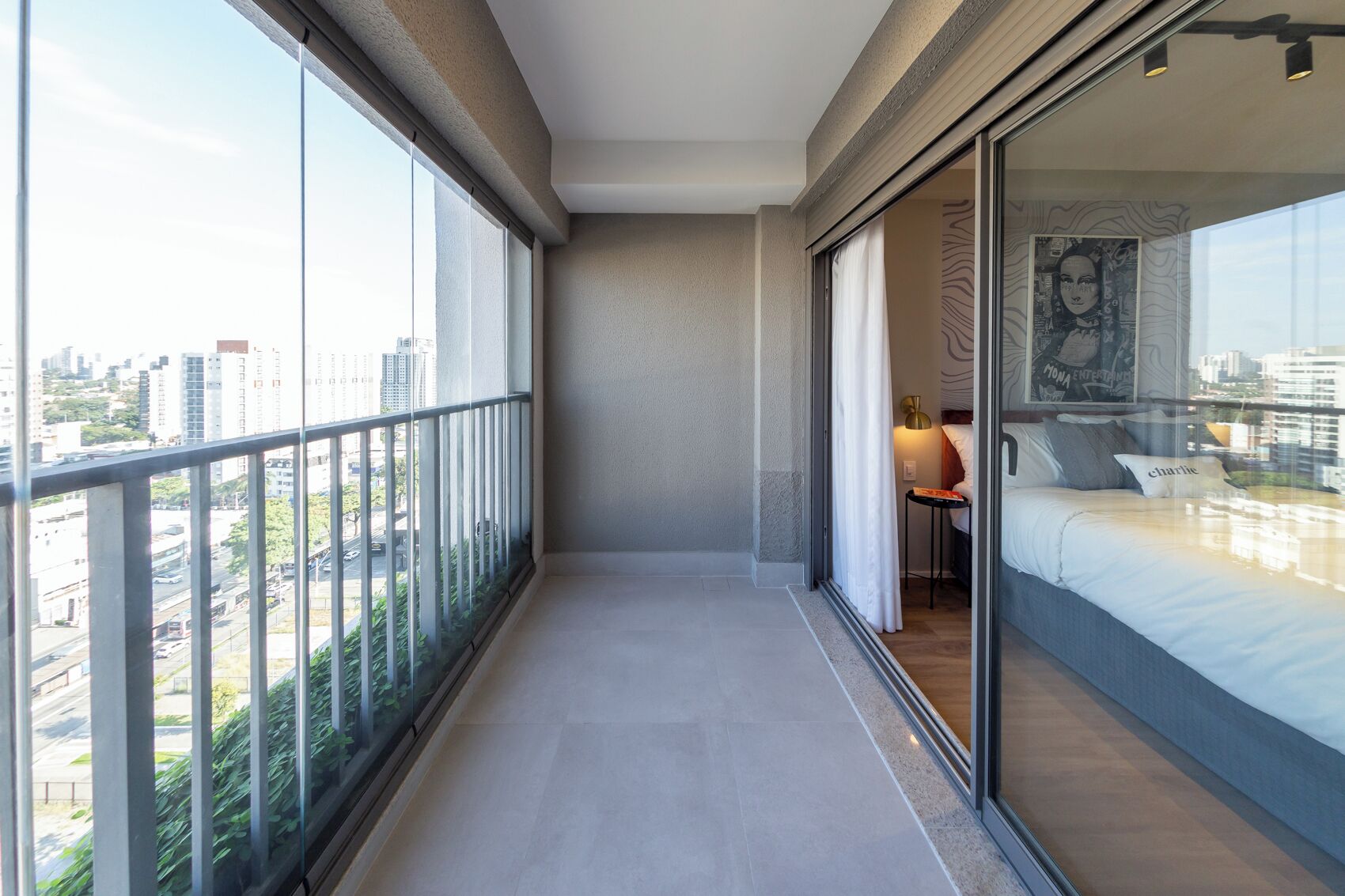 A narrow balcony with a city view, a bed with pillows, and a small table with a lamp.
