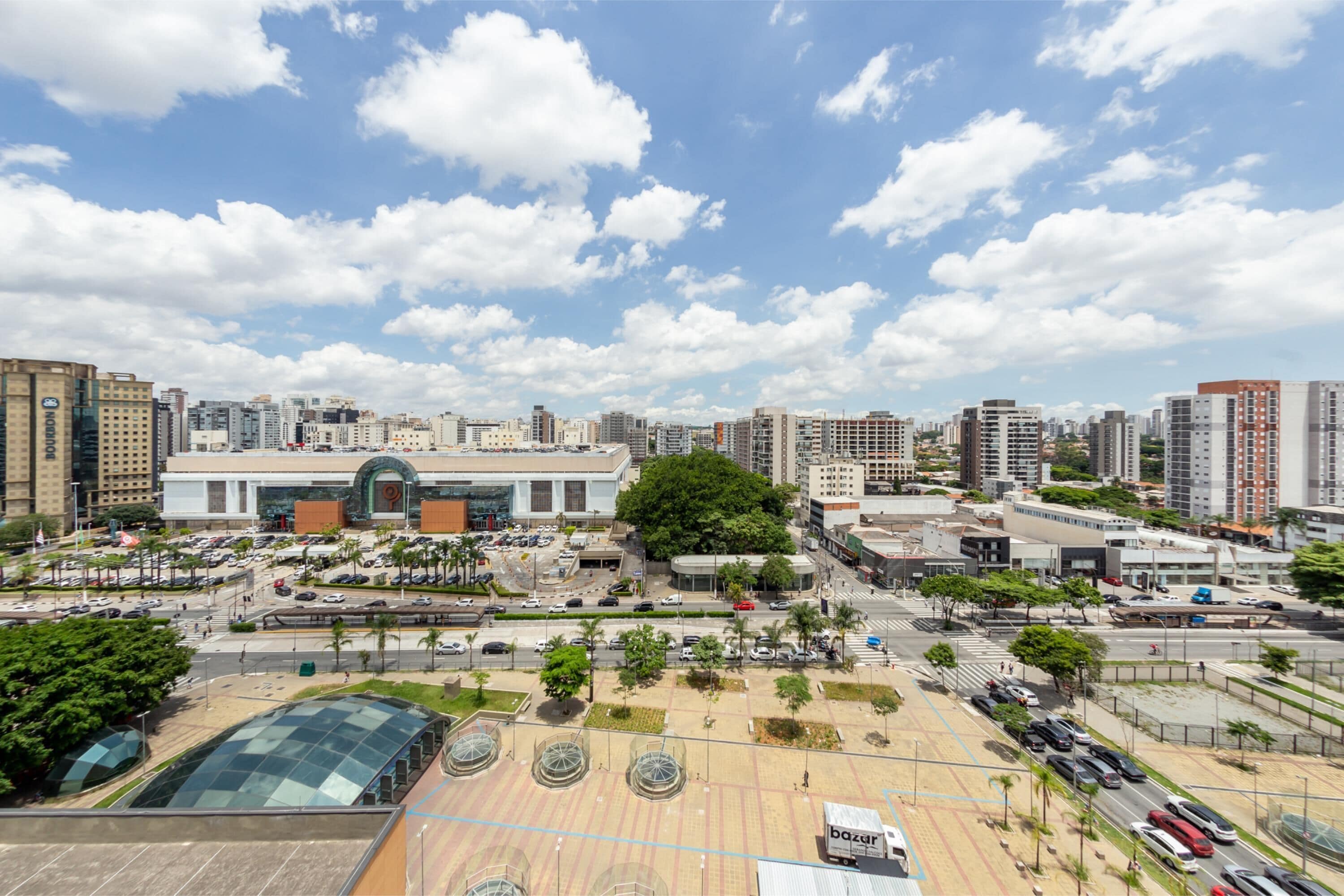 A cityscape with modern buildings, a large plaza with a glass structure, and a clear sky.
