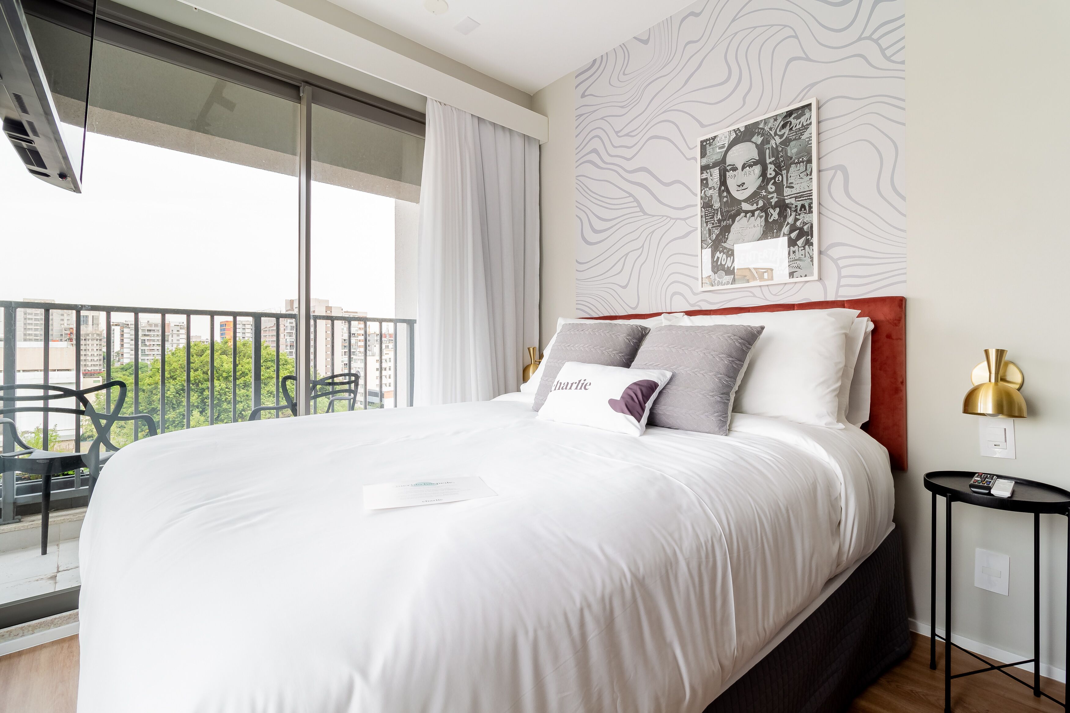 Ein Hotelzimmer mit einem großen Bett, Blick auf die Stadt durch raumhohe Fenster, einem modernen Wandbild und einem kleinen Nachttisch mit Lampe.