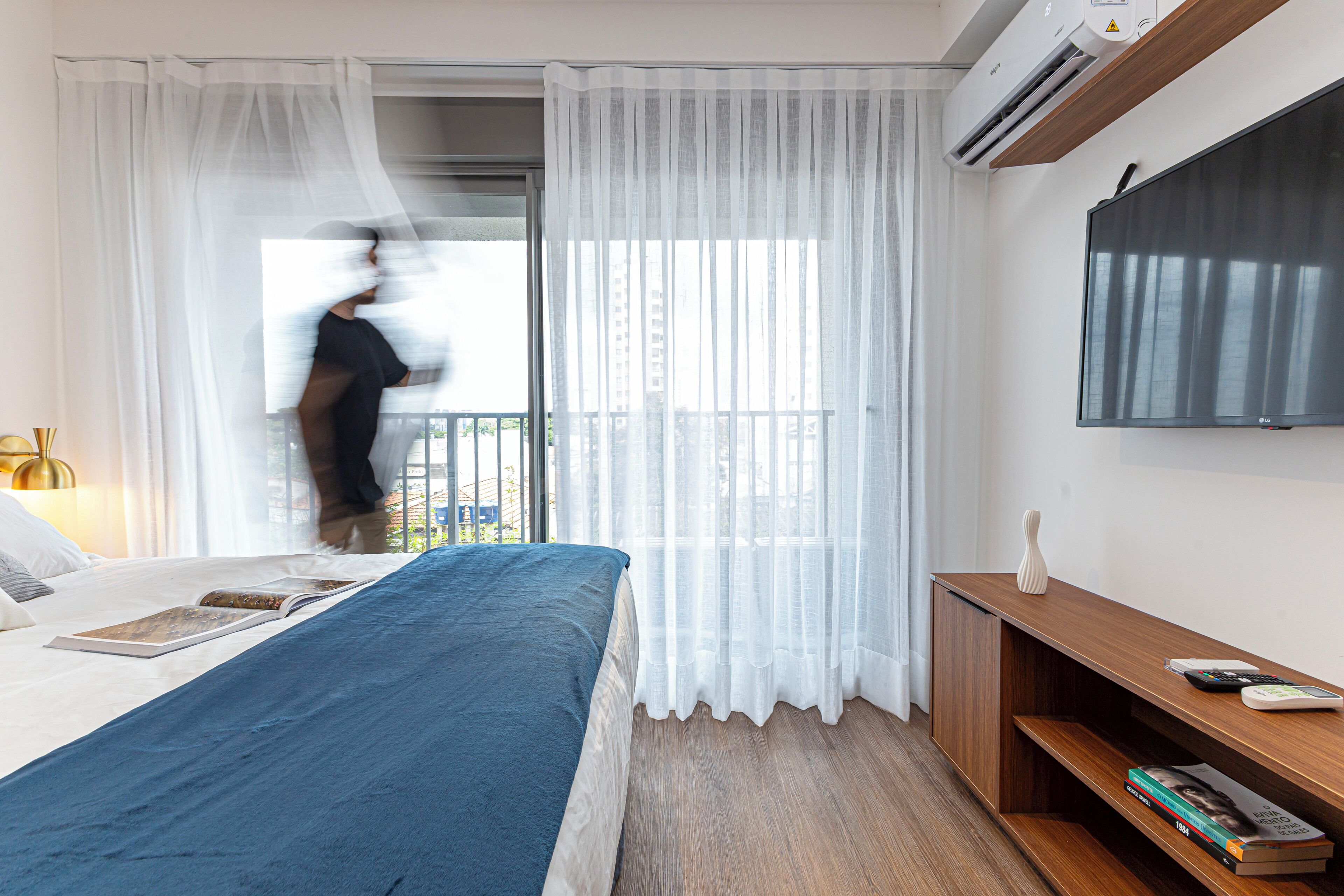 Una habitación de hotel con cama, televisor montado en la pared, una mesita de madera y cortinas traslúcidas.