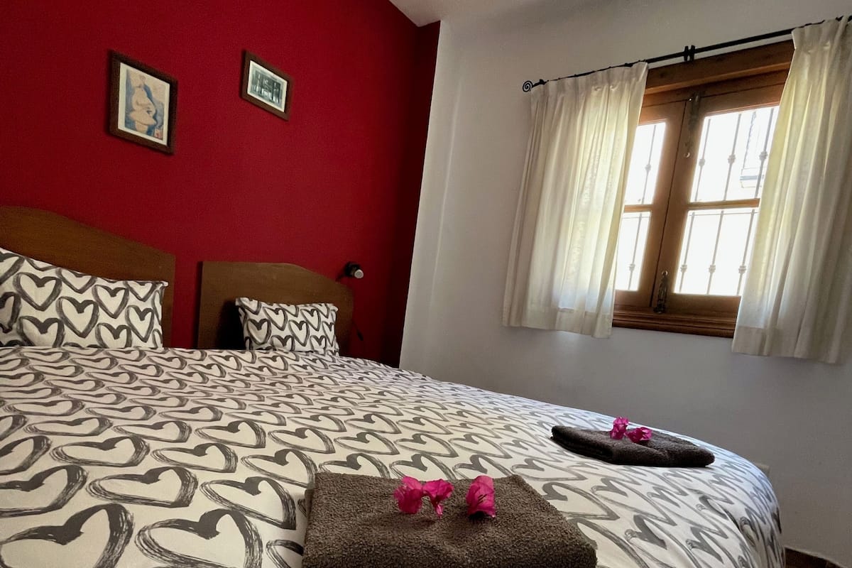 A hotel room with a large bed, red walls, a window with curtains, and two framed pictures on the wall.