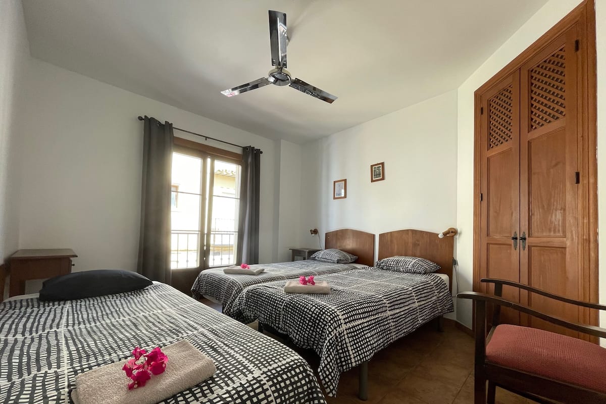 A hotel room with two beds, a ceiling fan, a wooden wardrobe, and a window with curtains.