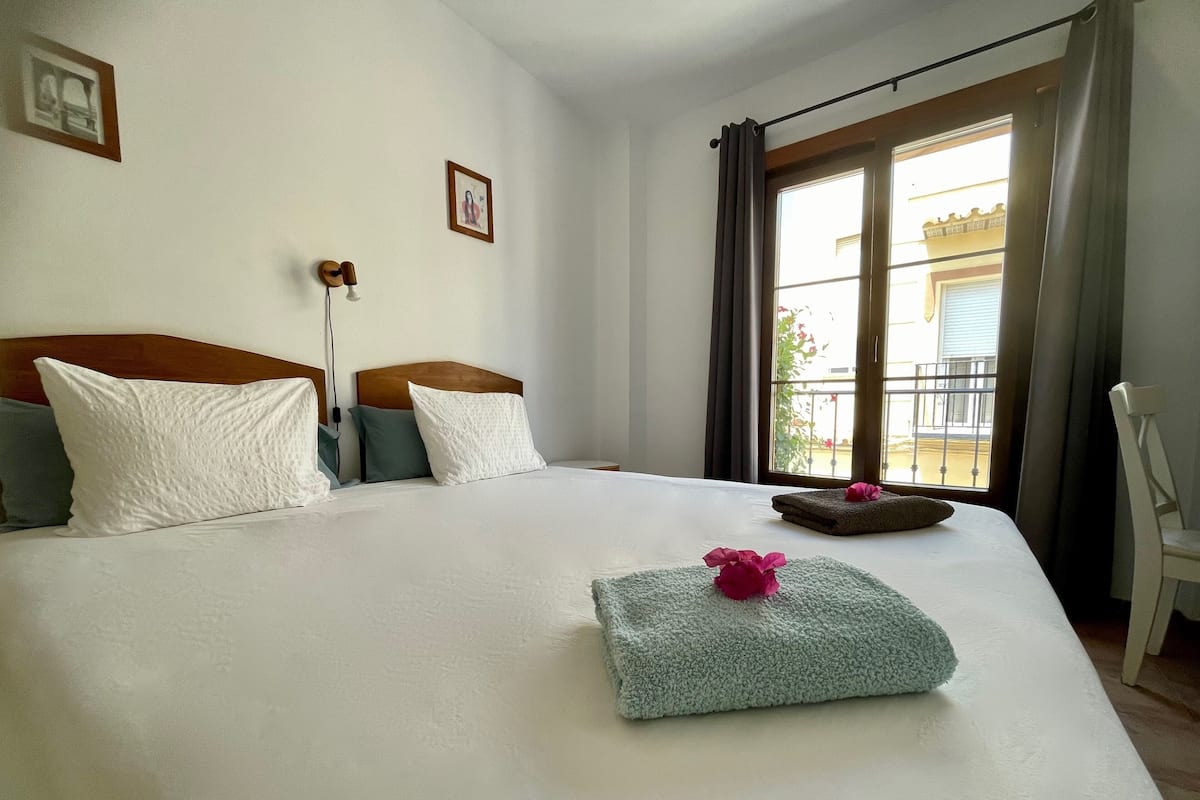 A hotel room with a large bed, a window with curtains, a chair, and a small table with a flower.